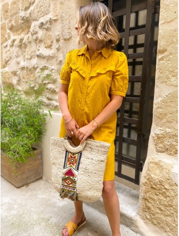 Femme portant une robe jaune en broderie anglais qui arrive au genou avec un sac de plage en osier.